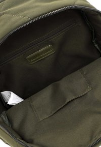 Interior de una mochila de tela verde oliva con un bolsillo con cremallera, textura suave y una etiqueta cosida; cuenta con un compartimento principal espacioso.