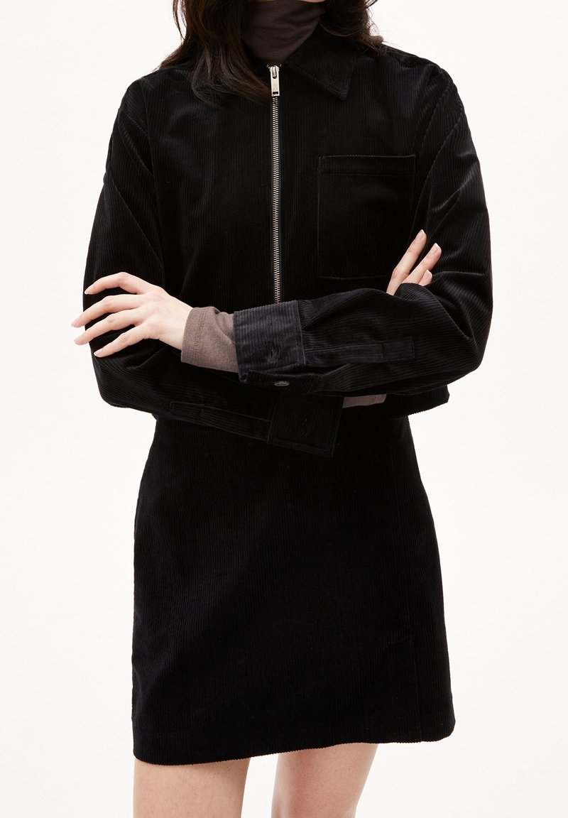 Czarna sukienka z korduroy z długimi rękawami, zamek błyskawiczny oraz kieszonką na piersi. Teksturowany materiał, dopasowany krój i prosty design.