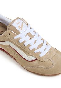 Zapatillas de ante beige con cordones blancos, detalles en rojo y blanco, perforaciones decorativas y un clásico logo de Vans en la lengüeta.