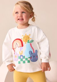 Chemise blanche à manches longues ornée d'un appliqué représentant un arc-en-ciel, un soleil, une poire, une fleur et un lapin bleu. Associée à des leggings jaunes.