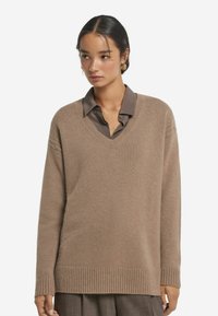 Beiger V-Ausschnitt-Pullover aus weichem Strick, mit gerippten Bündchen und Saum, über einem dunklen Hemd mit Kragen getragen, was einen geschichteten Look kreiert.