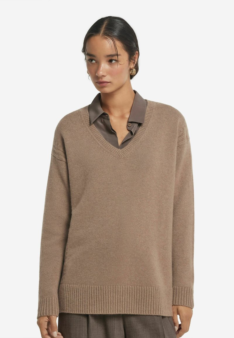 Beiger V-Ausschnitt-Pullover aus weichem Strick, mit gerippten Bündchen und Saum, über einem dunklen Hemd mit Kragen getragen, was einen geschichteten Look kreiert.