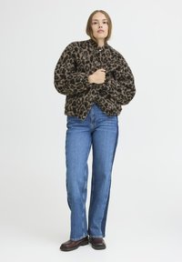 Veste en imprimé léopard brun et noir avec un col haut, des boutons pression et une texture duveteuse, associée à un jeans bleu avec un panneau latéral foncé.