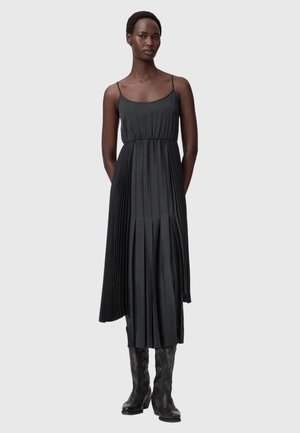 Kobieta ubrana w czarną plisowaną sukienkę midi na cienkich ramiączkach i czarne, sięgające do kolan haftowane botki, stojąca na jednolitym tle.