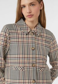 Žena v béžovém, hnědém a černém kostkovaném kabátu s velkými knoflíky a špičatým límcem, stojící mírně nalevo na bílem pozadí.