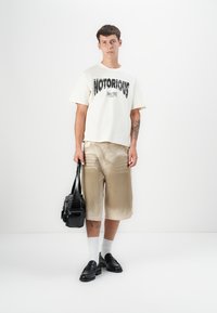 White T-shirt with "NOTORIOUS" print, beige shorts, black bag, and black shoes. Model wearing white socks, standing against a plain background.