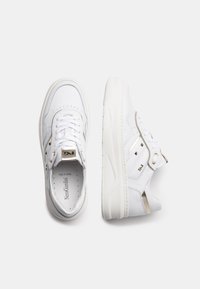 White sneaker with smooth leather upper, gold accents, six eyelets, and a thick, textured sole. Features a padded tongue and logo detail.