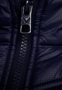 Marinblå pufferjacka med närbild av en svart dragkedja med en metallisk drare och texturerad söm längs kanten.