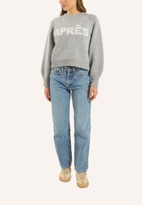 Grauer Strickpullover mit "APRES" in Weiß auf der Vorderseite, runder Ausschnitt und Ballonärmel, kombiniert mit hellblauen geraden Jeans und beigen Sneakern.