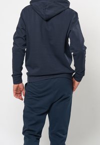 Marineblauer Hoodie mit einer abgerundeten Kapuze, gerippten Bündchen und einer weichen Textur, kombiniert mit passenden dunklen Hosen, von hinten betrachtet.