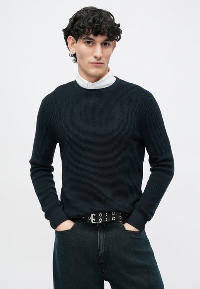 Jersey de punto marino con textura acanalada, cuello redondo y mangas ajustadas, combinado con denim oscuro y un cinturón negro con detalles circulares plateados.
