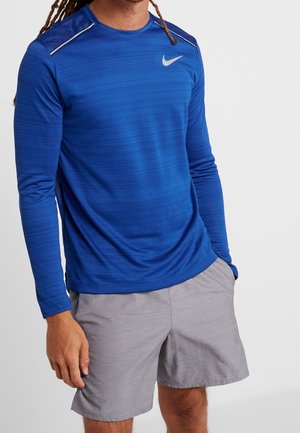 Vue recadrée d’un mannequin portant un haut de sport Nike bleu à manches longues et un short gris, sur un fond uni et clair.