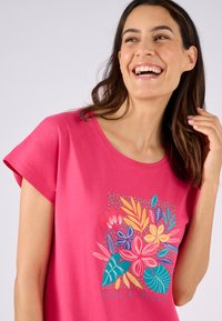 T-shirt di cotone rosa con maniche corte, caratterizzato da una stampa floreale colorata nelle tonalità di blu, giallo e rosso, etichettato "NOTTE TROPICALE".