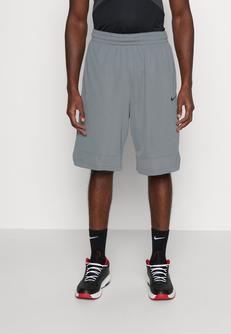 Homme portant un short Nike gris, des chaussettes Nike noires et des chaussures de sport noires avec des semelles rouges et blanches, debout devant un fond uni.
