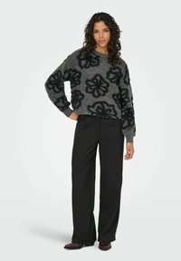 Frau steht mit der Hand auf der Hüfte, trägt einen grauen Pullover mit schwarzen Blumenmustern, schwarze weit geschnittene Hosen und dunkle Schuhe.