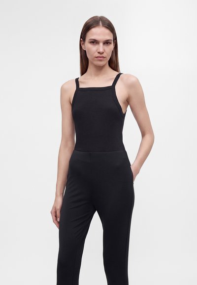 Jeune femme aux longs cheveux bruns portant un haut noir sans manches et un pantalon noir, debout avec une main dans sa poche devant un fond uni.