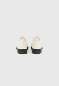 Zapatillas de lona blancas con suelas de goma negra y acentos texturizados, con un diseño de caña baja y una pestaña en la parte trasera.