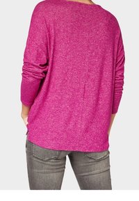 Kobieta w luźnym, długim, magentowym swetrze z długim rękawem, w połączeniu z szarymi dżinsami, pokazana z tyłu z rękami w kieszeniach.