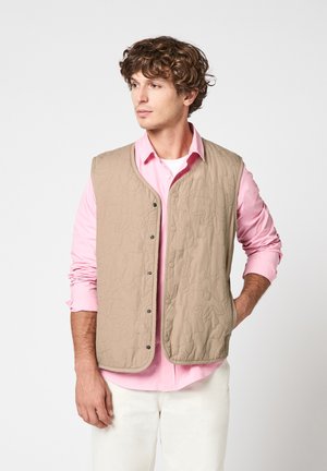 Jeune homme portant une veste matelassée beige sur une chemise rose et un pantalon blanc, debout avec une main dans la poche, regardant sur le côté sur un fond uni.