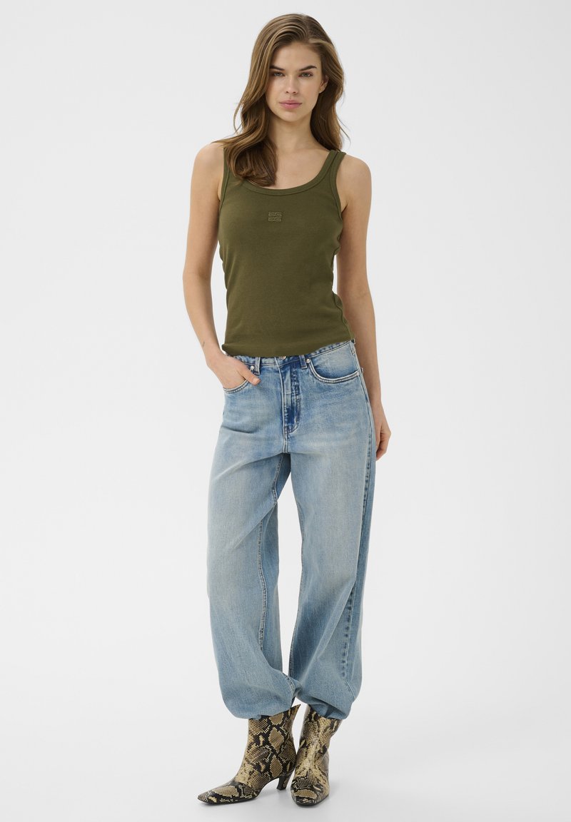 Femme portant un débardeur vert olive, un jean bleu clair ample et des bottines à imprimé serpent, debout avec une main dans la poche sur fond blanc.