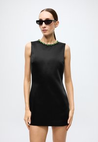 Black sleeveless mini dress with a smooth texture, featuring a green gem-accented neckline. Model wears narrow sunglasses.