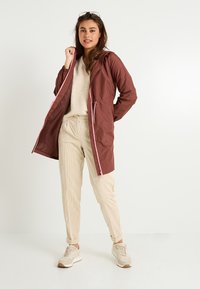 Veste imperméable marron avec une fermeture éclair rose, dotée d'une capuche et de poignets élastiques, portée sur un pull beige et un pantalon en velours côtelé.