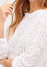 Frau mit langen roten Haaren, die eine weiße Spitzenbluse mit Puffärmel trägt und eine Haarsträhne nah an ihrem Gesicht hält.