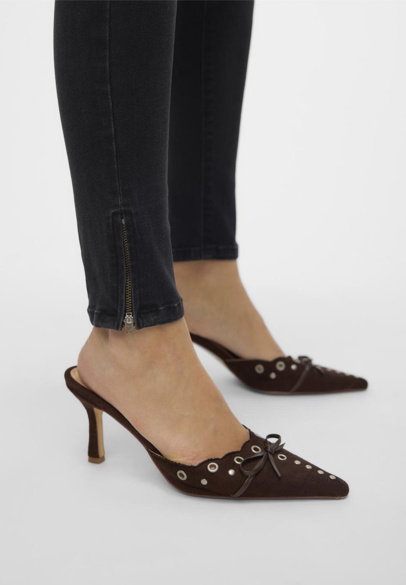Bruine suède muiltjes met puntige neuzen, golvende randen en zilveren oogjes. Voorzien van een klein strikje en een slanke hak.