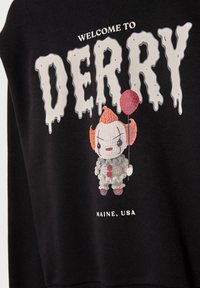 Indumento nero con testo "Benvenuti a Derry, Maine, USA" e un piccolo clown all'uncinetto che tiene un palloncino rosso.