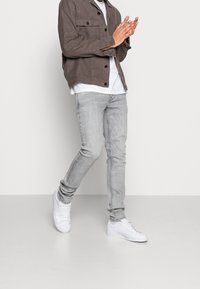 Persona vestida con jeans de mezclilla gris, zapatillas blancas, camiseta blanca y chaqueta marrón con botones, de pie y aplaudiendo.