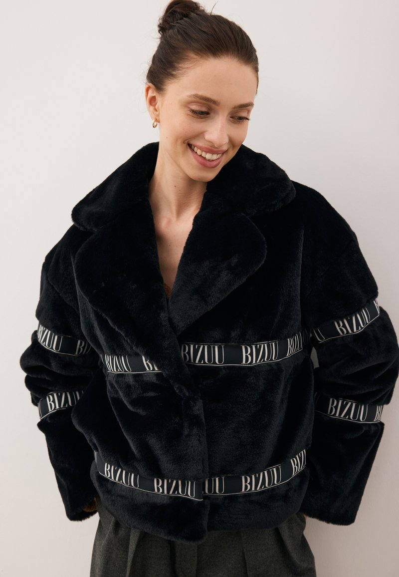 Czarna kurtka z sztucznego futra z kołnierzem szalowym, z szerokimi czarno-białymi pasami z napisem "BIZUU" na klatce piersiowej i rękawach.