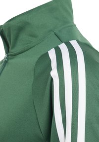 Zelena sportska jakna s visokim ovratnikom, zatvaranjem na zip i tri bijele pruge na ramenu. Tekstura materijala daje sportski izgled.
