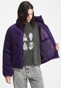 Paars corduroy pufferjack met ritssluiting, elastische manchetten en een glanzende voering, gedragen over een grafisch grijs t-shirt en versleten jeans.