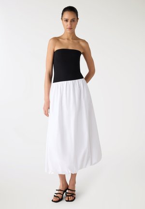 Donna con top nero senza spalline, gonna bianca a palloncino e sandali neri con cinturini, in piedi davanti a uno sfondo chiaro e semplice.