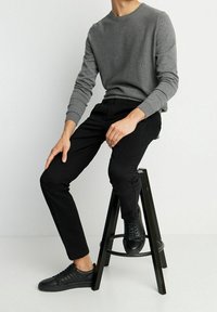 Szara dzianinowa bluzka z okrągłym dekoltem, w połączeniu z czarnymi spodniami typu slim fit i czarnymi skórzanymi sneakersami, siedząca na czarnym stołku.