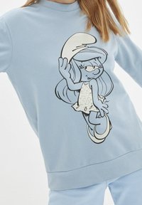 Niña vistiendo un sudadera azul claro con un personaje de dibujos animados, Smurfette, sosteniendo su sombrero y sonriendo.