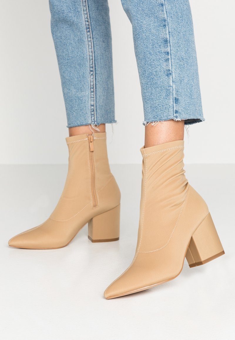 Bottines beiges à bout pointu avec talons blocs et fermetures éclair latérales portées avec un jean court bleu clair sur un sol blanc.