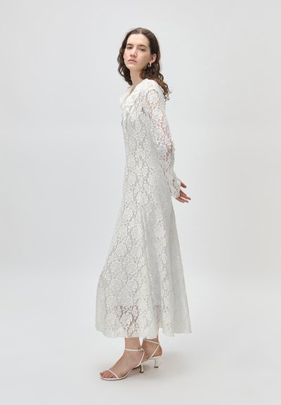 Νεαρή γυναίκα στέκεται φορώντας ένα μακρύ λευκό δαντελένιο φόρεμα με μακριά μανίκια και λευκές φιάπες με λουράκια, σε απλό ανοιχτόχρωμο φόντο.