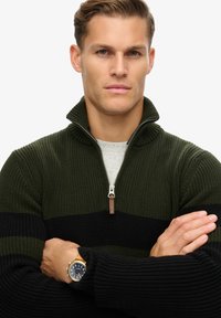Zielono-czarny sweter z zapięciem na zamek, wykonany z dzianiny, z żeberkową strukturą, z srebrnym zamkiem błyskawicznym, noszony na jasnoszarym koszuli.