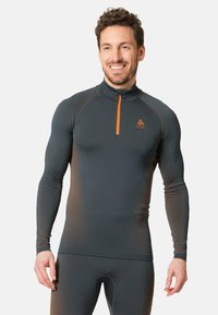 Sportieve long-sleeve top in donkergrijs met een oranje rits en logo. Beschikt over textiel met een structuur en een gestippeld verlooppatroon op de mouwen.