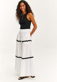 Weißer, mehrlagiger Maxirock mit schwarzen Rüschenakzenten, kombiniert mit einem enganliegenden schwarzen ärmellosen Oberteil und schwarzen Sandalen. Weicher Stoff, lockere Passform.