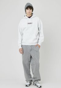 Sweat à capuche gris avec cordons, doté d'une poche avant et de texte brodé. Associé à un jean gris décontracté et à un bonnet gris. Baskets blanches avec des accents noirs.