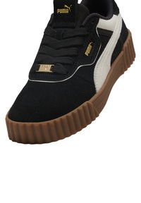 Zapatillas de ante negro con detalles en blanco, suela de goma marrón texturizada, diseño de cordones y detalle de logo dorado en la lengüeta y el lado.