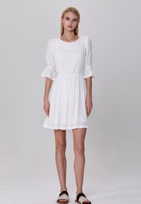 Robe blanche courte en tissu texturé, à encolure ronde et manches volantes. Présente des bordures en dentelle sur les manches et une taille cintrée.