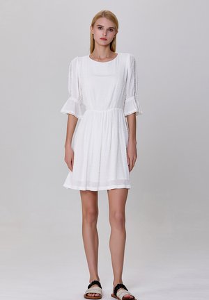 Robe blanche courte en tissu texturé, à encolure ronde et manches volantes. Présente des bordures en dentelle sur les manches et une taille cintrée.