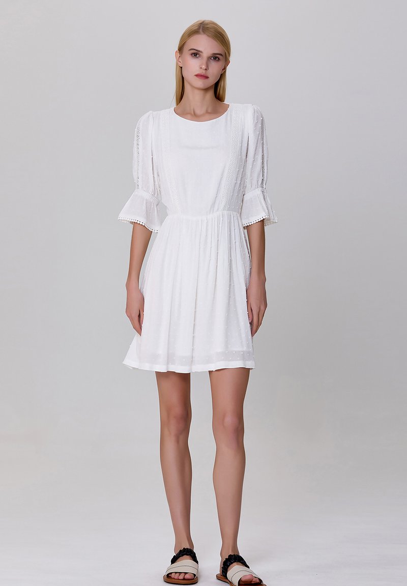 Robe blanche courte en tissu texturé, à encolure ronde et manches volantes. Présente des bordures en dentelle sur les manches et une taille cintrée.