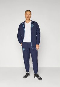Donkerblauw trainingspak met lichtblauwe accenten. Bestaat uit een jas met rits en logo, en bijpassende broek. De broek heeft elastische manchetten en zakken.