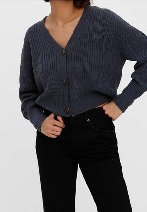 Femme portant un cardigan en maille épaisse gris foncé boutonné avec col en V et un pantalon noir taille haute, une main dans la poche.