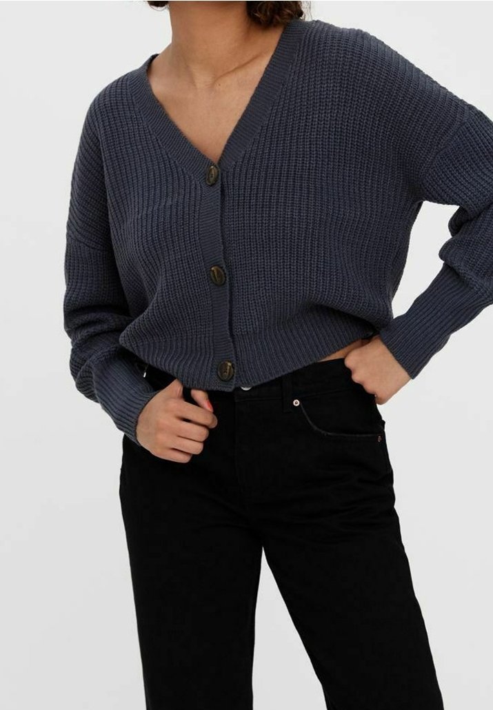 Femme portant un cardigan en maille épaisse gris foncé boutonné avec col en V et un pantalon noir taille haute, une main dans la poche.