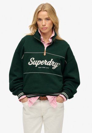 Pull-under vert en polaire avec zip quart, logo et rayures blanches, superposé sur une chemise à rayures roses, associé à un pantalon crème.